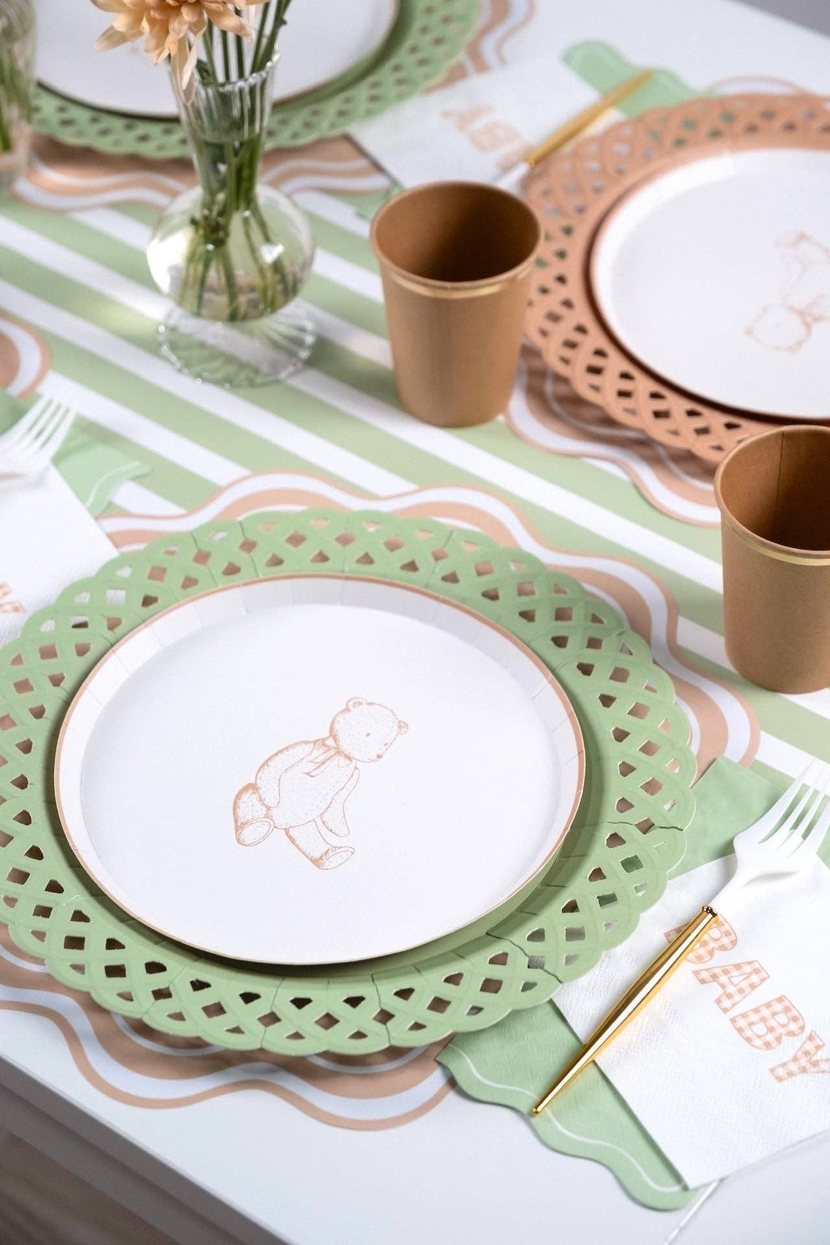 Place setting close-up showing a bunny plate on a green scalloped charger with the sage napkin, tan cups, and a green-striped runner.