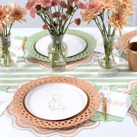 Styled place setting showing a bunny plate on a tan scalloped charger with the sage napkin, fresh flowers in vases, and a green-striped runner.
