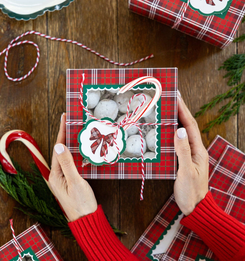 Red plaid Christmas cookie box with window filled with snowball cookies, tied with striped twine and a bow gift tag; candy canes, greenery, and plaid bow plate shown separately, not included