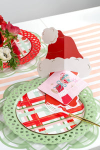 Place setting with a green lattice charger, red-and-white plaid plate, a folded red Santa-hat centerpiece, a pink present-print towel, and fresh flowers.