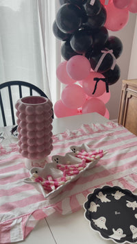 Happy Halloween felt pennant flag with a ghost design and striped ribbon tails, a white ghost-shaped divided tray filled with pink and white candy, a pink scalloped vase with ghost-print ribbon, a pink and white striped ruffled runner, and a black and pink balloon garland