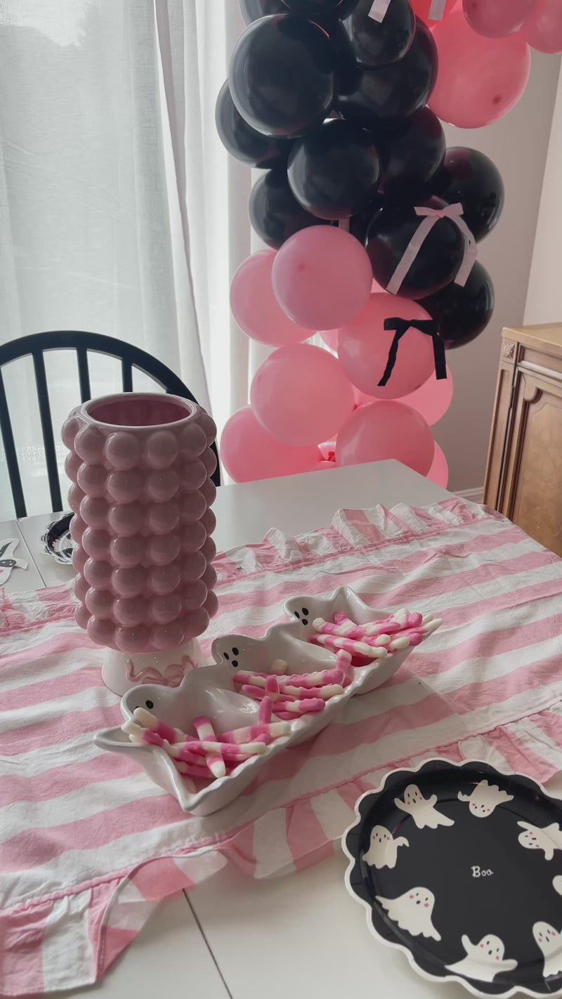 Happy Halloween felt pennant flag with a ghost design and striped ribbon tails, a white ghost-shaped divided tray filled with pink and white candy, a pink scalloped vase with ghost-print ribbon, a pink and white striped ruffled runner, and a black and pink balloon garland