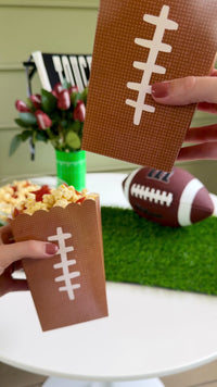 Football party snack setup with football-shaped treat boxes, popcorn, football roses, decorative banner, and football décor.