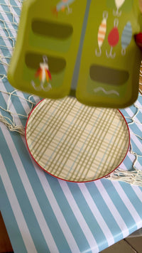 Fishing birthday party table setting featuring a vest-shaped plate, plaid dinner plate, scalloped placemat, striped cup, fish napkin, and tackle box.