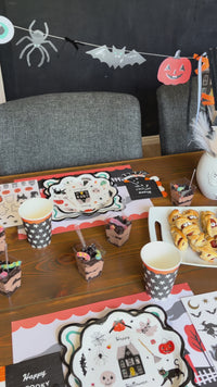 Happy Halloween scalloped paper plate with a black scalloped edge and cream ground, scattered with Halloween icons—haunted house, black cat, pumpkin, ghost with a BOO sign, witch hat, spider, and winged eyeball—shown in a kids' Halloween party setting