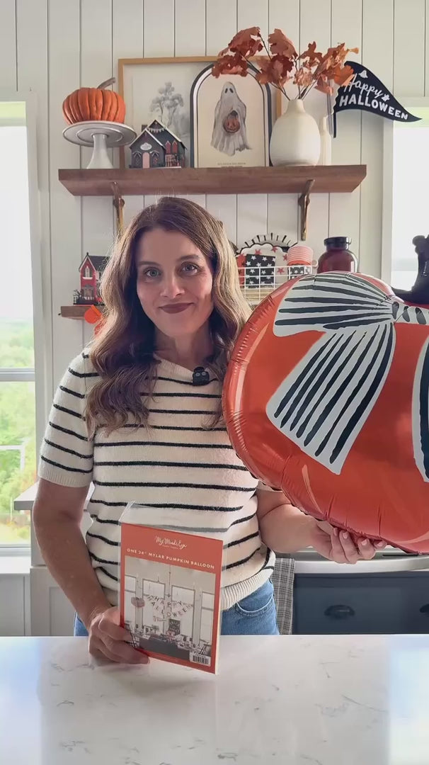 a woman holding the inflated pumpkin bow balloon in a styled Halloween kitchen with a garland and haunted house décor.