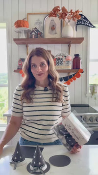 a woman styling the witch hat treat holders on a kitchen counter with a Halloween garland, jars, and fall décor.