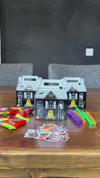 Assembled haunted house paper treat boxes with a black mansion facade, shingled roof, glowing yellow door, ghost, bat, spiderweb, and pumpkin, each with a cut-out carry handle, styled on a Halloween dessert table with cupcakes, candy-corn-edged napkins, a Stay Spooky pennant, and balloons