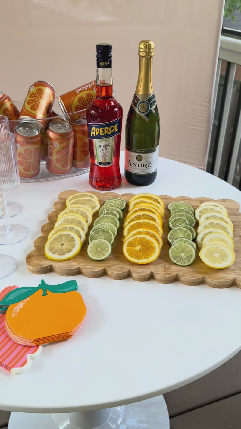 A bright and modern summer drink station styled on a round white table. The setup features ingredients for a citrus cocktail or mocktail, including a bottle of Aperol, André Champagne, an ice bucket of orange sodas, ribbed fluted glassware, and a beautifully arranged tray of sliced lemons, limes, and oranges.