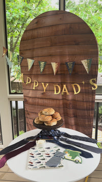 Video walkthrough of a Father's Day party table styled on a screened porch, featuring megaphone print paper dinner plates, blue striped tie-shaped napkins, and golf cart shaped napkins; pans across neckties used as table props, a stacked bagel display on a dark wood cake stand, and a wood-grain arch backdrop hung with a gold "Happy Dad's Day" letter banner and multicolor pennant flag garland