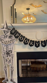 Slow pan of a Halloween mantel with a black "Spooky Season" pennant banner, gold paper bats flying across the mirror, a skull and gold pumpkins, black taper candles, and jointed paper skeletons in witch hats on either side of the fireplace.