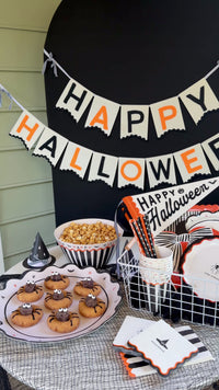 Halloween party table styled with a cream felt Happy Halloween banner, spider cookies, caramel popcorn, and black and orange tableware