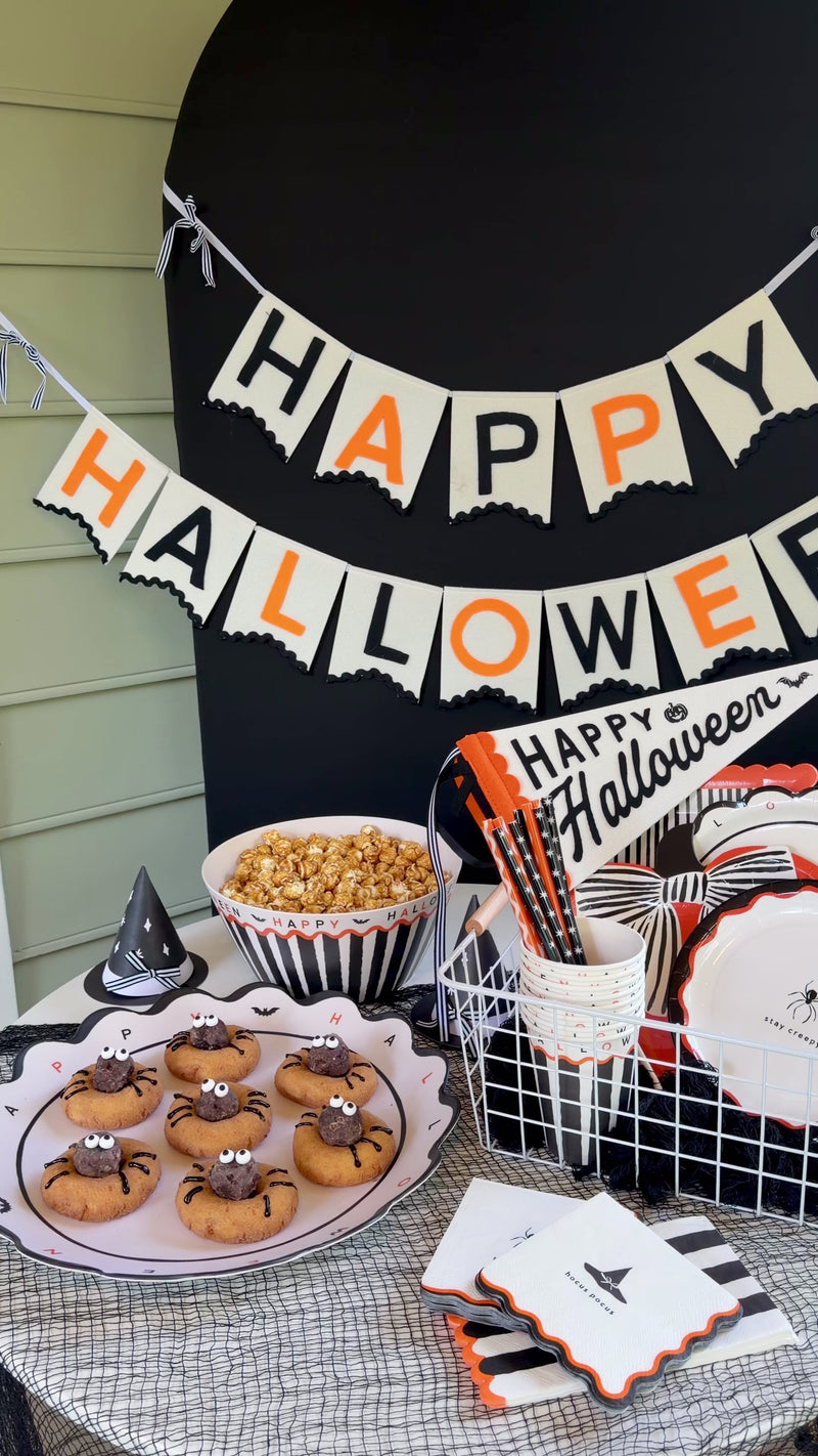 Halloween party table styled with a cream felt Happy Halloween banner, spider cookies, caramel popcorn, and black and orange tableware