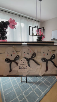 Ghost felt banner from the Booquette collection with 10 soft white felt ghosts and black bows strung across a 5-foot garland, styled as pink Halloween decor along a shelf and table in a black, white, and pink room