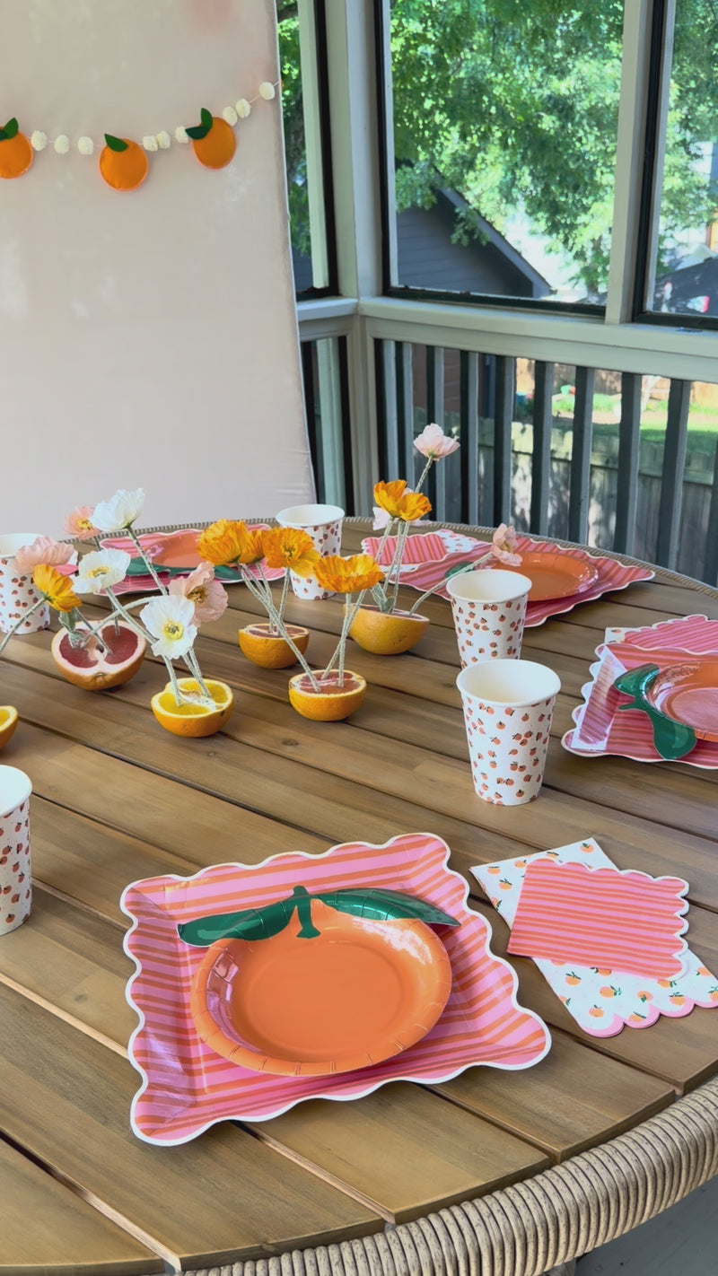 A vibrant lifestyle video featuring an orange and citrus-themed summer party table setting. Includes unique centerpieces made of crepe-paper flowers inside real halved oranges and grapefruits, paired with layered pink striped and orange-shaped plates, fruit-print cups, and a matching felt garland