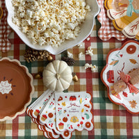 Pumpkin pie paper plate with a scalloped crust edge beside a white pumpkin, bowl of popcorn and Happy Thanksgiving napkins on a fall plaid tablecloth