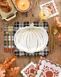 A styled autumn table setting featuring a white pumpkin shaped plate with a gold pumpkin outline, a black-and-white plaid napkin, gold flatware, patterned napkins, and fall leaves on a rustic wooden table.