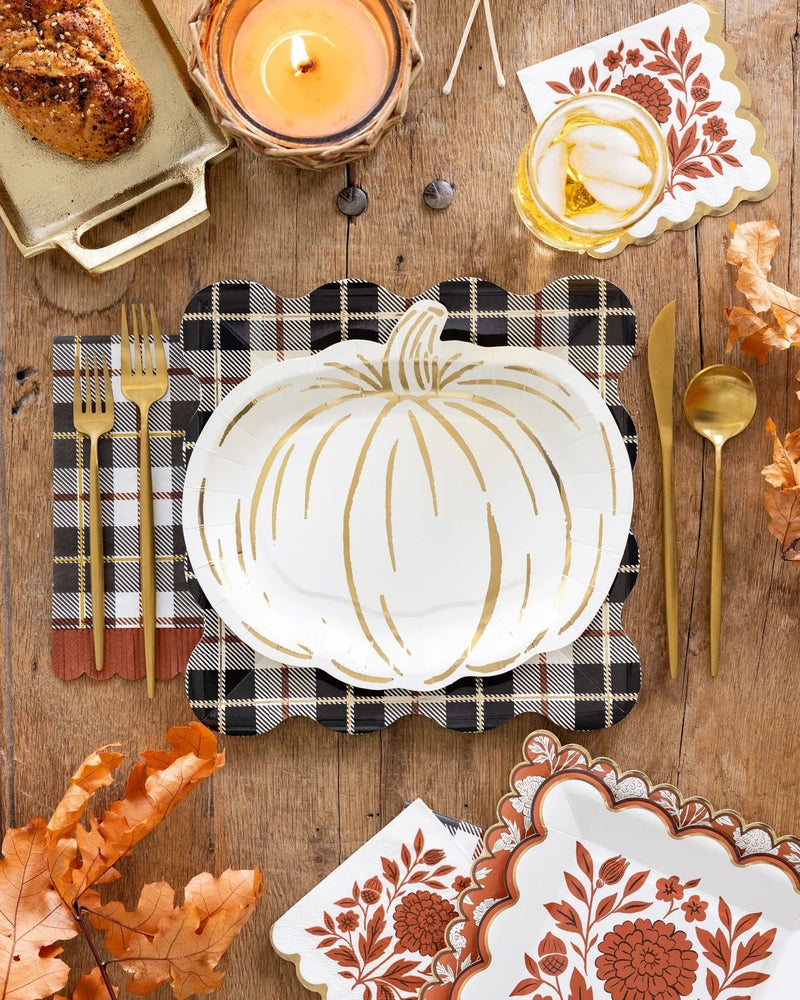A styled autumn table setting featuring a white pumpkin shaped plate with a gold pumpkin outline, a black-and-white plaid napkin, gold flatware, patterned napkins, and fall leaves on a rustic wooden table.