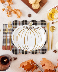 A styled autumn table setting featuring a white pumpkin-shaped plate with gold accents, placed on a black and white plaid napkin with gold flatware. The scene is decorated with orange fall leaves, star anise, a candle, cookies on a wooden board, and a glass of iced tea on a light beige surface.