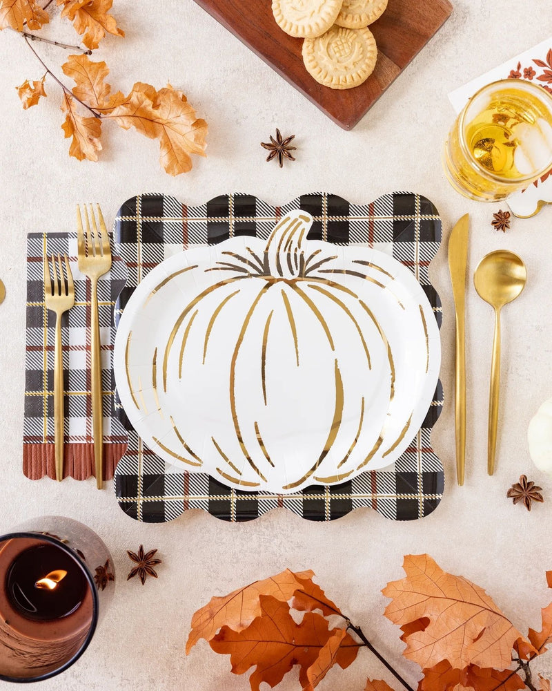 A styled autumn table setting featuring a white pumpkin-shaped plate with gold accents, placed on a black and white plaid napkin with gold flatware. The scene is decorated with orange fall leaves, star anise, a candle, cookies on a wooden board, and a glass of iced tea on a light beige surface.