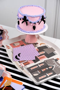 Stack of purple checkered bat napkins on a haunted house tray beside a pink and lavender Halloween cake