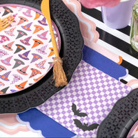 Close view of a lavender bat cocktail napkin beside a scalloped black plate rim and broom stirrer