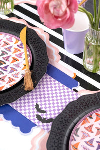 Purple checkered bat napkin tucked between a black scalloped plate and a black and white striped runner