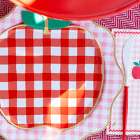 Red gingham apple shaped paper plate set as a place setting on a pink gingham charger with a matching apple napkin and red-handled knife.