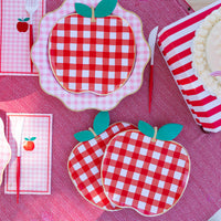 Overhead picnic table setting with red gingham apple shaped paper plates, pink gingham napkins, red cutlery, striped cups, and a layer cake.