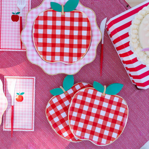 Overhead picnic table setting with red gingham apple shaped paper plates, pink gingham napkins, red cutlery, striped cups, and a layer cake.