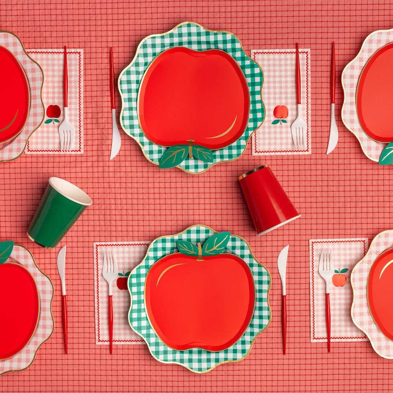 Overhead tablescape with red apple shaped paper plates layered on top of green gingham scalloped charger plates, styled with apple-print napkins, red and green paper party cups, red and white striped cutlery, and a red gingham table runner