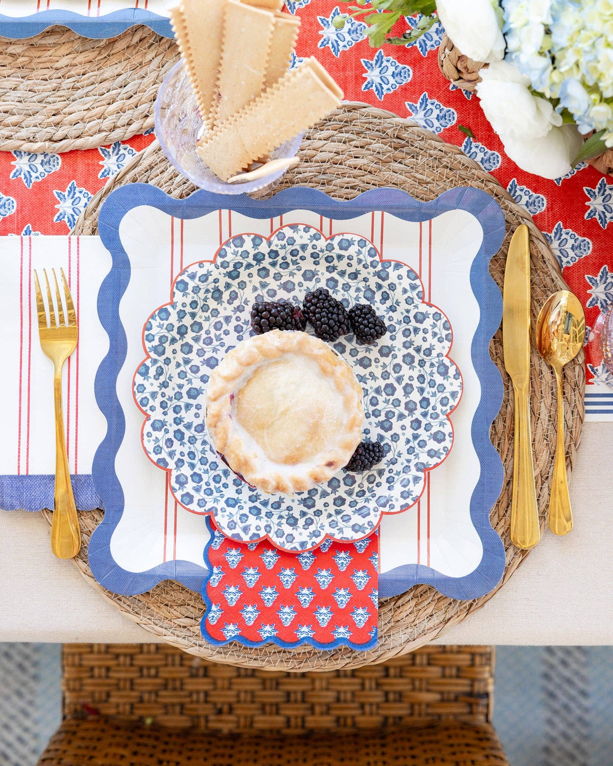 Red block-print floral paper napkin layered with blue scalloped dinner plate, floral salad plate, mini pie, and gold cutlery on wicker placemat with red and blue Hamptons table setting