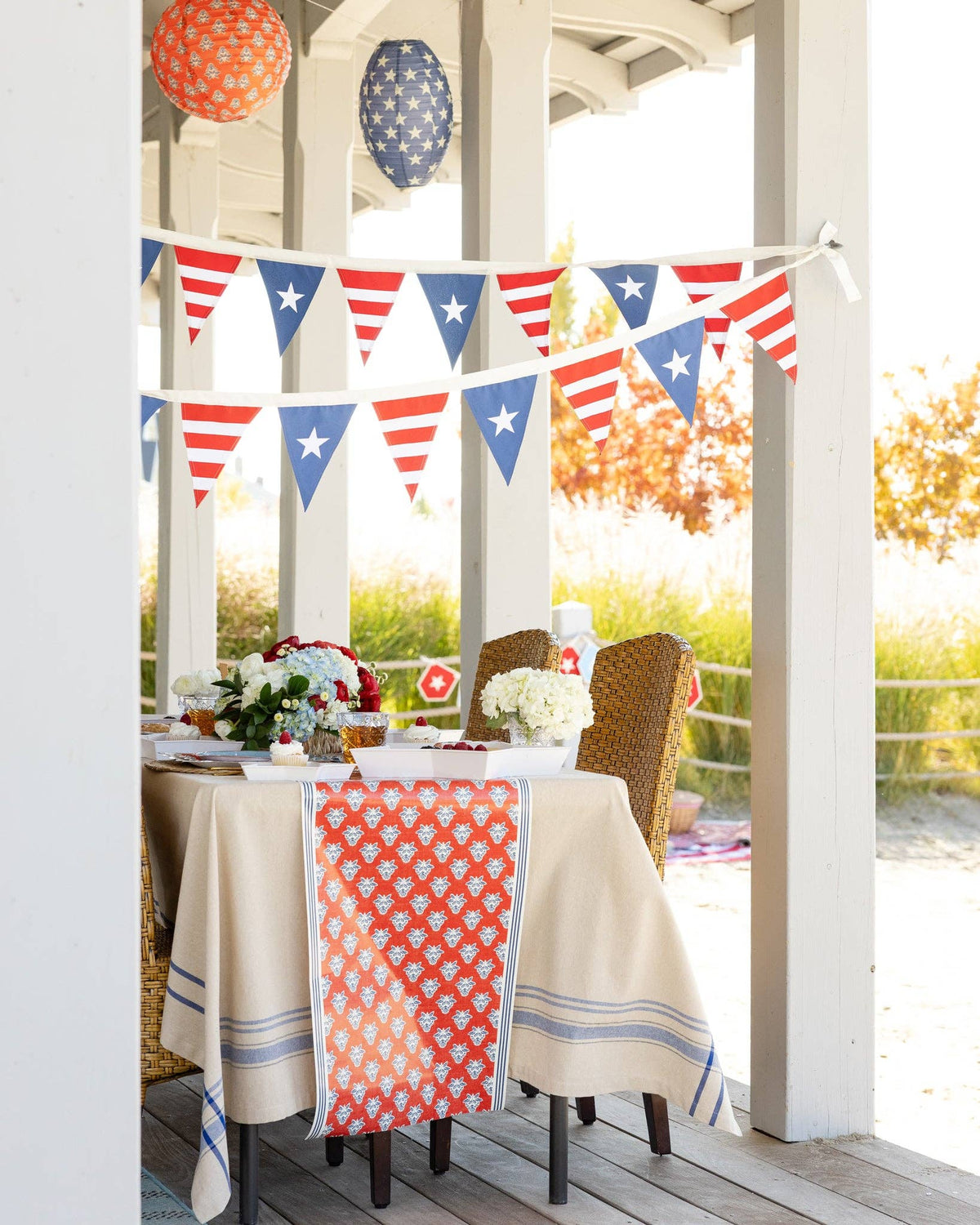 Porch dinner table with red block print floral paper table runner draped over linen tablecloth with patriotic pennant bunting overhead