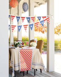 Porch dinner table with red block print floral paper table runner draped over linen tablecloth with patriotic pennant bunting overhead