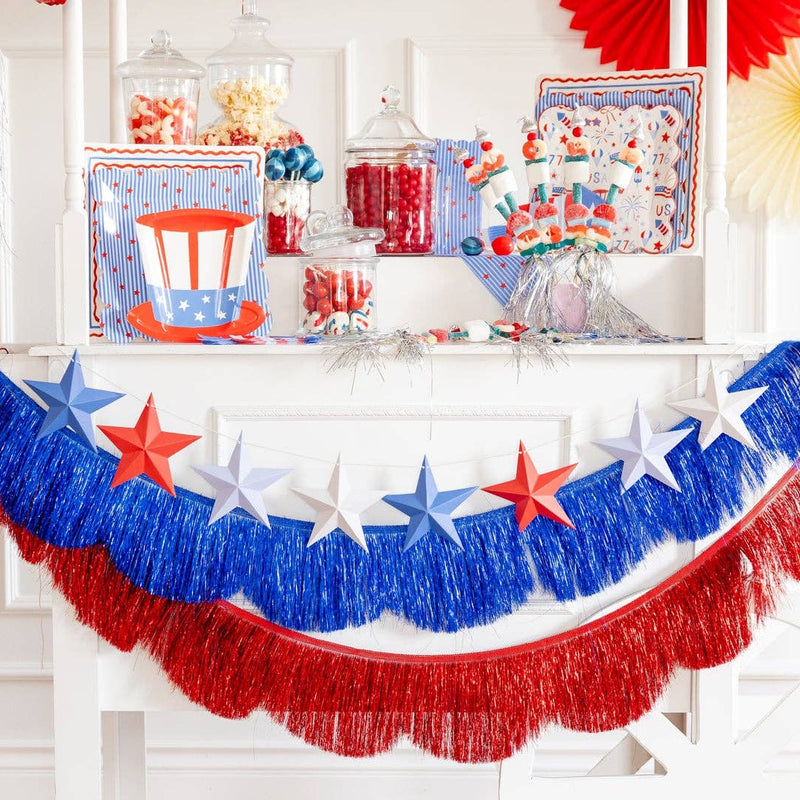 Close-up of blue and red metallic mylar fringe banners layered together in a swag on a white party cart with a 3D paper star garland and patriotic tableware in the background