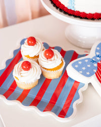 Overhead close-up of cherry-topped cupcakes on a red and blue striped scalloped paper plate next to a bow shaped napkin and cake stand