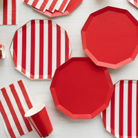 Solid red and red striped paper plates with red and white striped guest towel napkins and red paper cups on a white wood table.