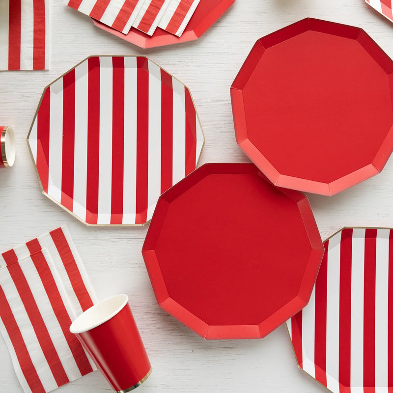 Solid red and red striped paper plates with red and white striped guest towel napkins and red paper cups on a white wood table.