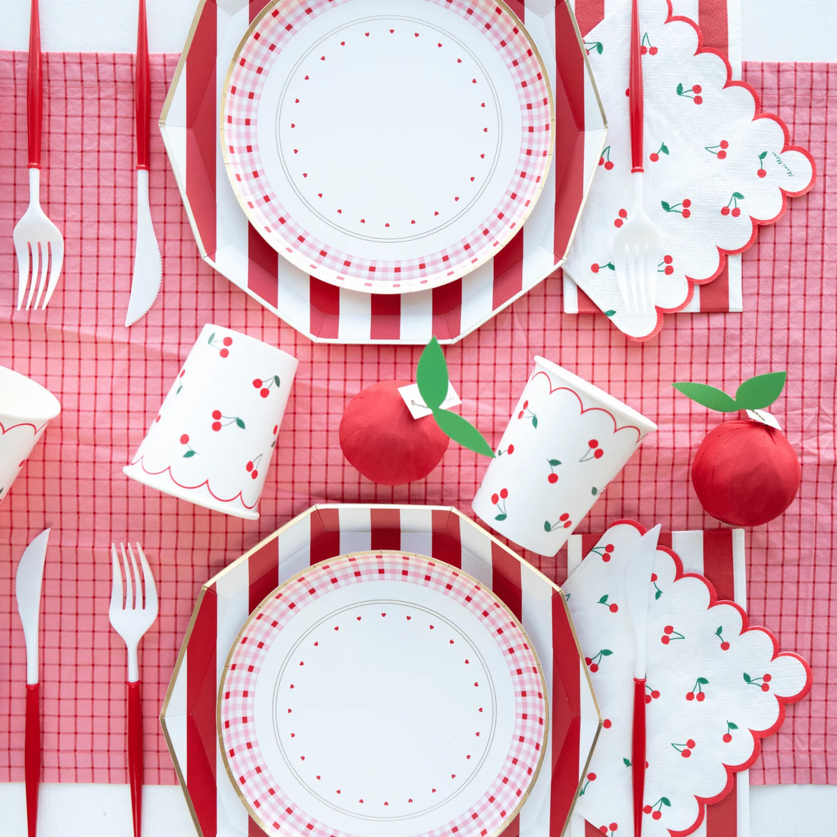 Cherry party table setting with red and white cabana stripe dinner plates, pink gingham plates, cherry print cups, and scalloped cherry napkins on a red grid table cover.
