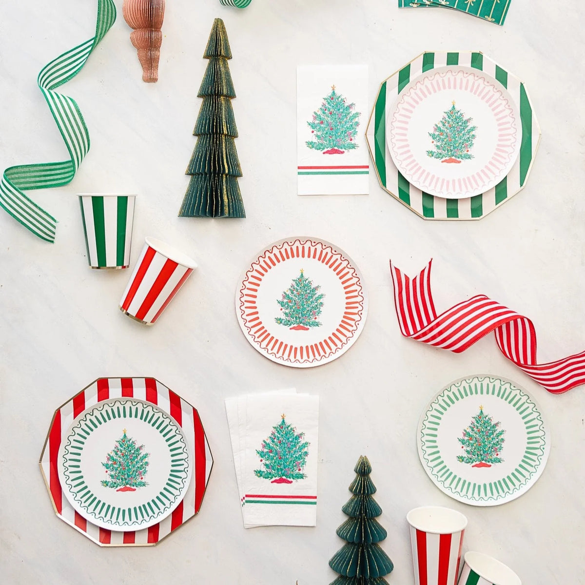 Christmas tree paper plates layered on green and red cabana stripe dinner plates with striped paper cups, tree napkins, and paper honeycomb trees on a white surface.