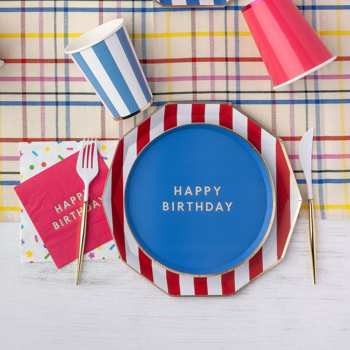 Blue Happy Birthday paper plate layered on a red and white cabana stripe dinner plate with striped paper cups, pink birthday napkins, and gold flatware on a plaid tablecloth.
