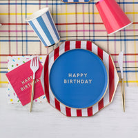 Blue Happy Birthday paper plate layered on a red and white cabana stripe dinner plate with striped paper cups, pink birthday napkins, and gold flatware on a plaid tablecloth.