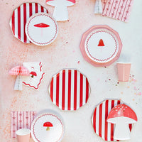 Mushroom paper plates and napkins layered on red and white cabana stripe dinner plates with pink bow napkins and blush paper cups on a speckled pink background.