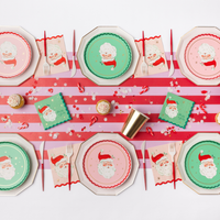 Red-and-green Christmas tablescape with green and pink Santa plates, the Santa napkins, gold cups, and a striped candy-cane runner.