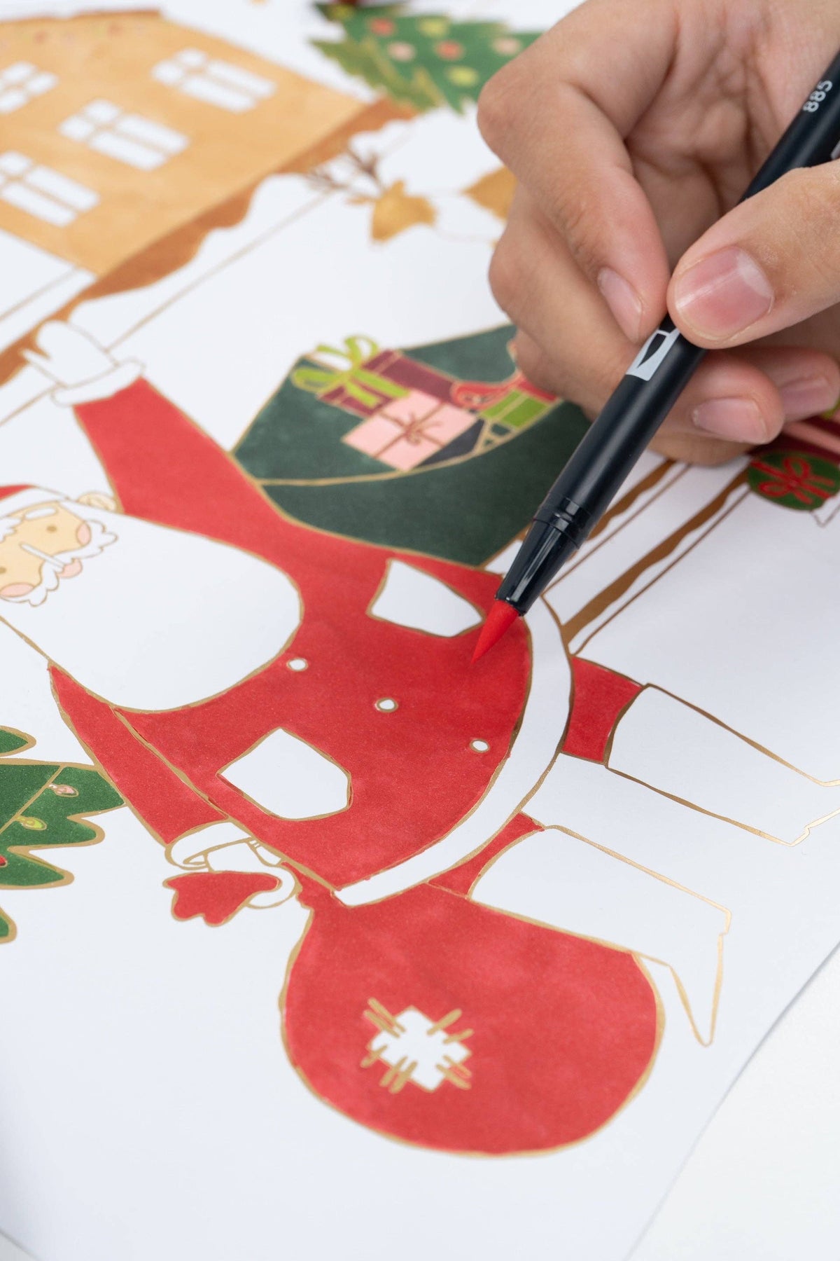Macro detail of a hand using a red marker to fill in Santa's suit, highlighting the shiny gold foil borders.