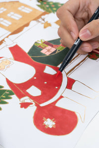 Macro detail of a hand using a red marker to fill in Santa's suit, highlighting the shiny gold foil borders.