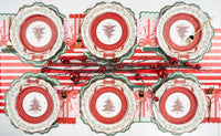 Overhead of several settings with red-medallion Christmas-tree plates, green scalloped chargers, a striped runner, and red bells.