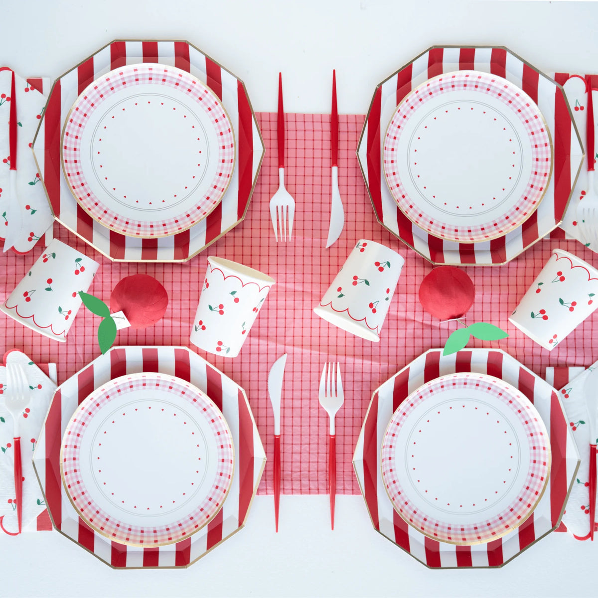 Cherry party table setting with red and white cabana stripe dinner plates, pink gingham plates, cherry print cups, and scalloped cherry napkins on a red grid table cover.