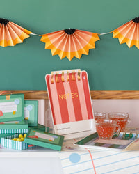 Red and pink striped notebook shaped paper plate displayed upright on an acrylic stand against a green chalkboard wall with pencil shaving accordion banner and school-themed treat boxes