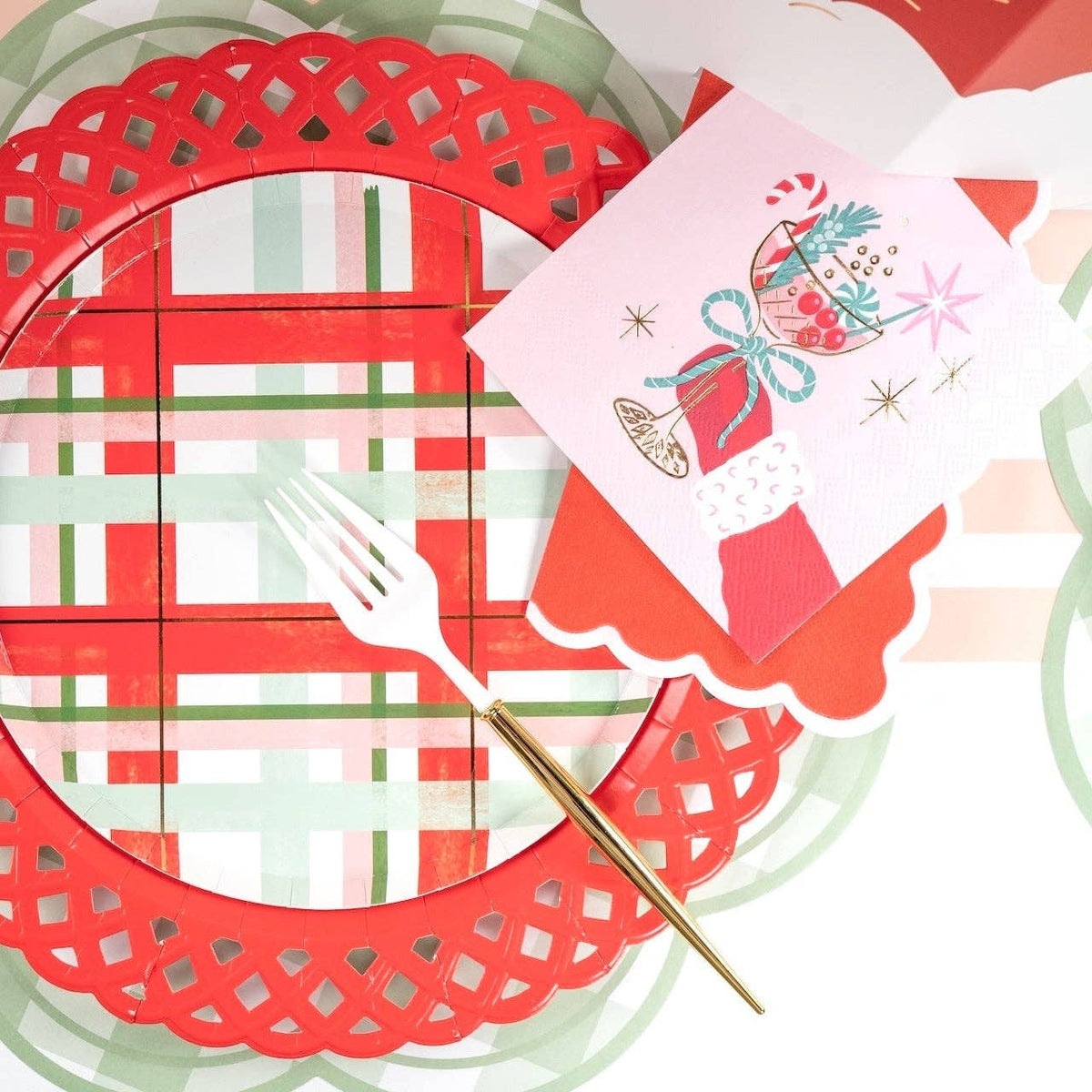 Overhead view of a festive place setting featuring a red die-cut scalloped charger plate, a plaid round dinner plate, and the pink retro cocktail napkin.